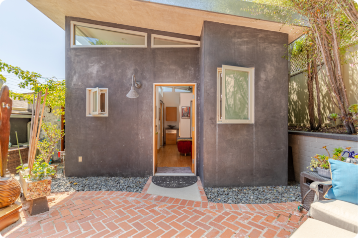 Tiny house confortable et moderne dotée d'une façade sombre, de grandes fenêtres et d'une porte d'entrée ouverte donnant sur un intérieur lumineux, entourée d'une terrasse en briques et d'une végétation luxuriante.