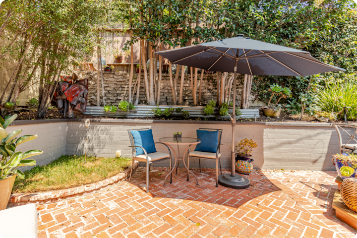 Patio extérieur confortable par une journée ensoleillée, avec deux chaises et une table sous un parasol noir, entourés de verdure et de plantes en pot.