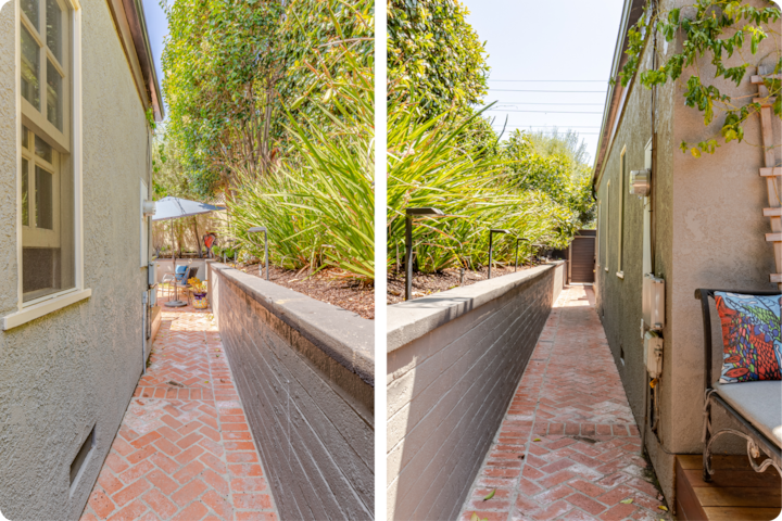 Un étroit chemin en briques bordé d'une plate-bande surélevée avec des plantes vertes luxuriantes sépare deux bâtiments beiges sous un ciel dégagé.