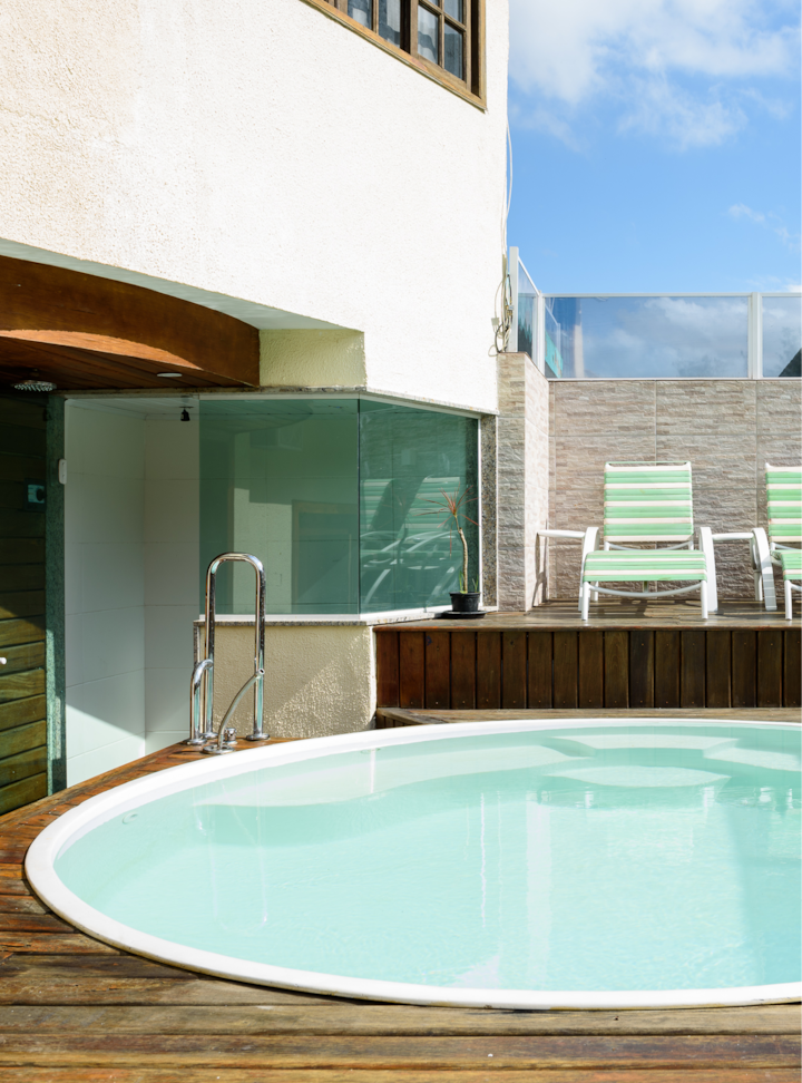 Piscine extérieure à l'eau transparente entourée d'une terrasse en bois, d'une balustrade en verre et de chaises longues à rayures vertes sous un ciel bleu.