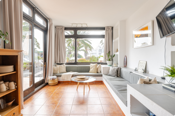Salon lumineux et confortable avec de grandes fenêtres donnant sur des palmiers, un canapé gris intégré, une table basse ronde en bois et un sol en terre cuite.