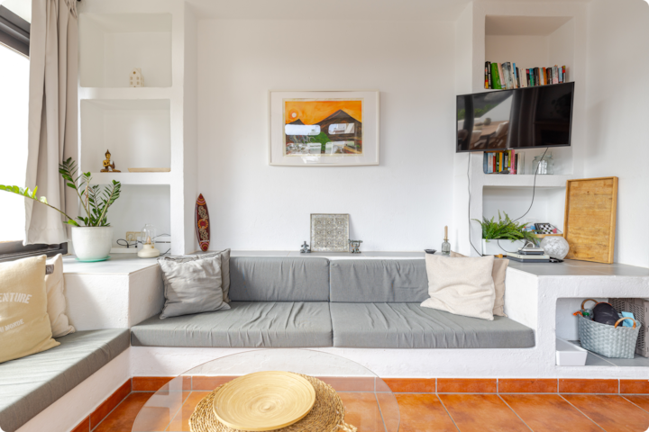 Salon confortable avec des sièges rembourrés gris, des étagères intégrées présentant des livres et des objets de décoration, et une télévision murale dans un espace lumineux et minimaliste. Un tableau représentant un paysage est suspendu au-dessus des sièges.