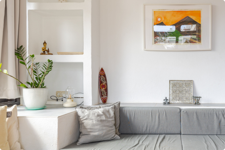 Un salon blanc confortable avec une étagère intégrée sur laquelle se trouvent une petite statue de Bouddha, une plante en pot et une lanterne, des sièges gris avec des coussins et un tableau abstrait sous cadre accroché au mur, représentant un coucher de soleil orange vif et des maisons.