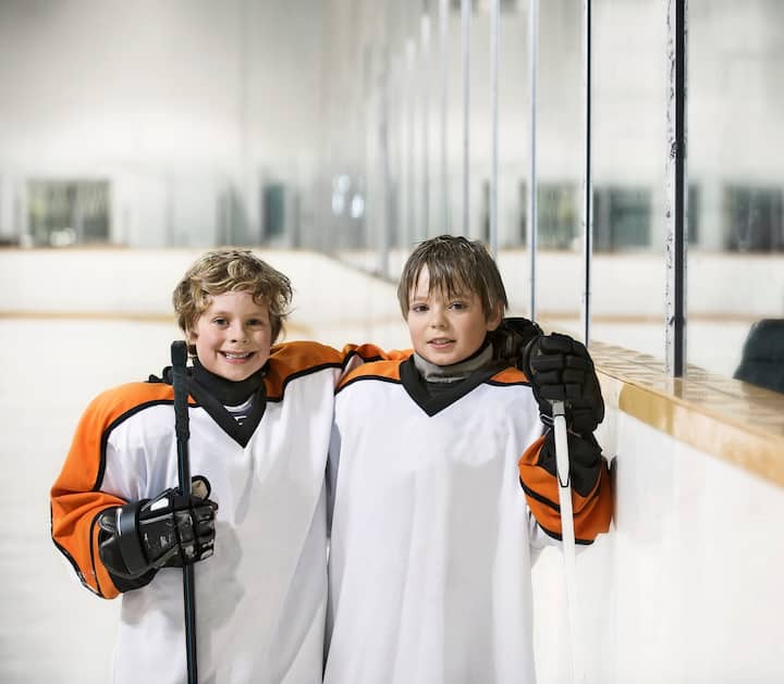 Two youth hockey players in white and orange uniforms hold hockey sticks and smile while standing on an indoor ice rink.