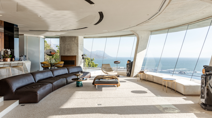 Salon moderne et spacieux avec une baie vitrée incurvée offrant une vue panoramique sur l'océan, doté d'un élégant canapé modulable en cuir noir et d'un mobilier minimaliste, baigné d'une lumière naturelle qui inonde l'espace.