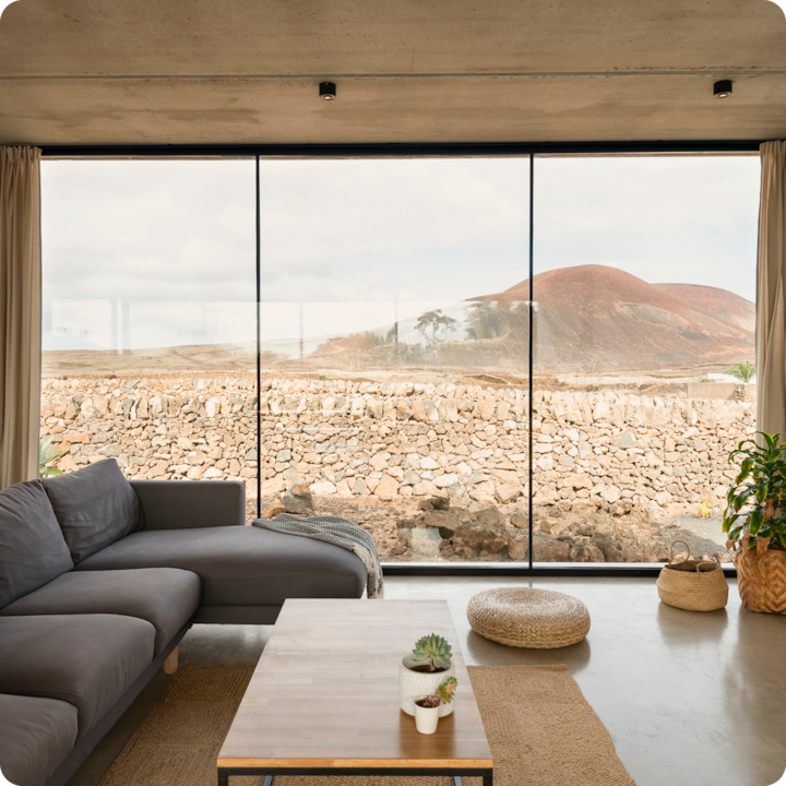 Un salon moderne avec un canapé gris, une table basse en bois sur laquelle sont posées de petites plantes en pot et une grande baie vitrée offrant une vue sur un paysage rocheux et une colline rouge et arrondie sous une lumière douce et diffuse.