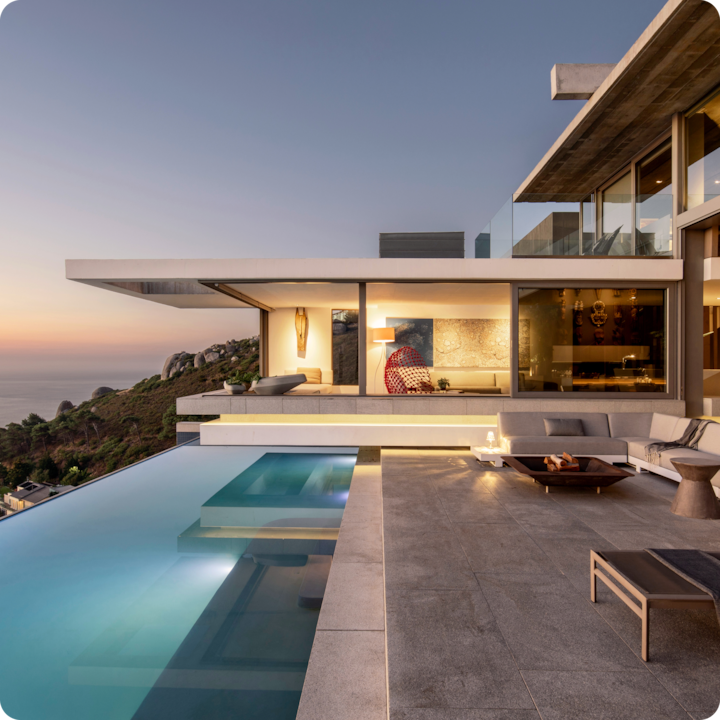Maison moderne à flanc de colline au coucher du soleil avec une piscine à débordement surplombant l'océan, dotée d'un patio spacieux et d'un éclairage intérieur chaleureux.