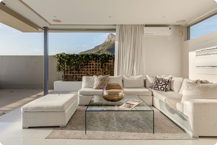 Un salon lumineux et moderne avec un canapé modulable blanc, une table basse en verre sur laquelle se trouvent un vase en verre et des magazines, et des portes coulissantes donnant sur un patio avec vue sur une montagne et sur de la verdure.