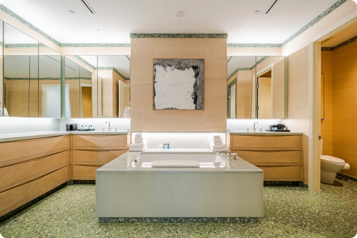 Salle de bain moderne avec une grande baignoire centrale, du carrelage vert au sol et des meubles en bois, entourée de miroirs et d'un éclairage doux.