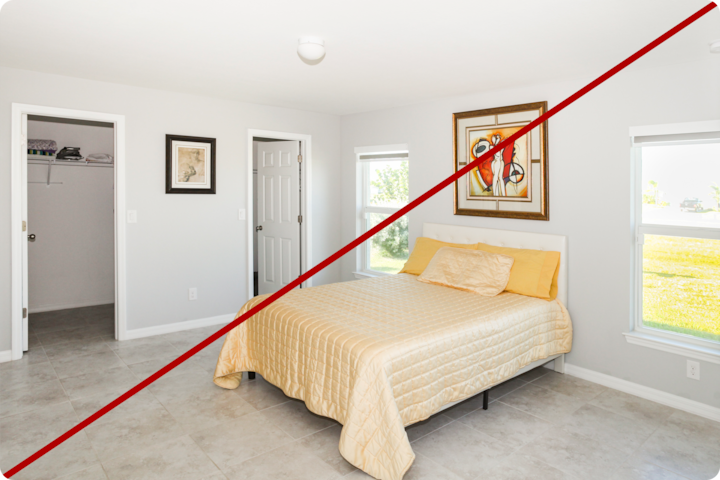 Une chambre minimaliste avec un lit soigneusement fait, de la literie jaune, deux grandes fenêtres laissant entrer la lumière naturelle, un tableau abstrait encadré au-dessus du lit et un petit dressing dans le coin de la pièce. Une ligne diagonale rouge traverse l'image, indiquant qu'il s'agit d'une photo prise avant des améliorations ou d'un exemple à ne pas suivre.