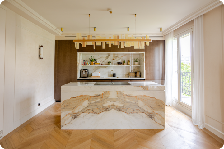 Une cuisine moderne avec un îlot en marbre aux veines beiges et dorées symétriques, des étagères ouvertes dotées de plantes et d'appareils de cuisine, et de grandes fenêtres laissant entrer la lumière naturelle.