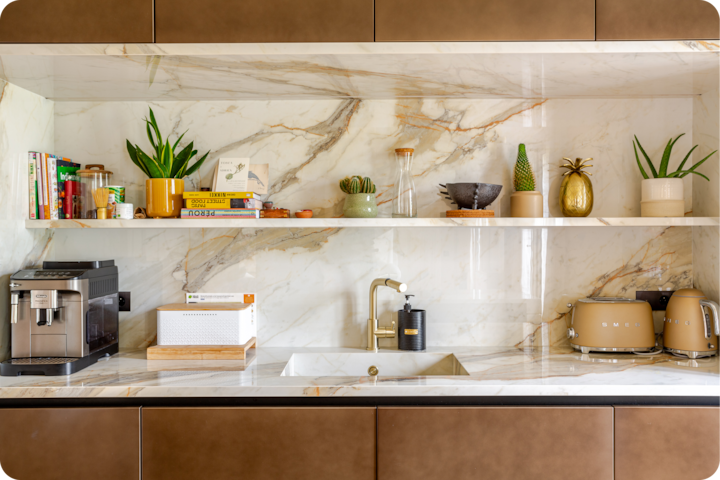 Plan de travail d'une cuisine moderne avec crédence en marbre, robinet doré et étagères ouvertes dotées de plantes, de livres et d'objets de décoration, à côté d'une machine à café et d'appareils électroménagers beiges.