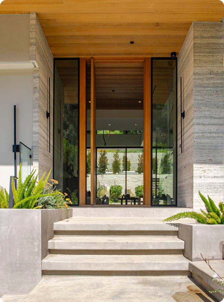 Une entrée moderne avec des marches en béton, des portes en bois, des parois en verre et une végétation luxuriante des deux côtés, menant à un espace intérieur ouvert.