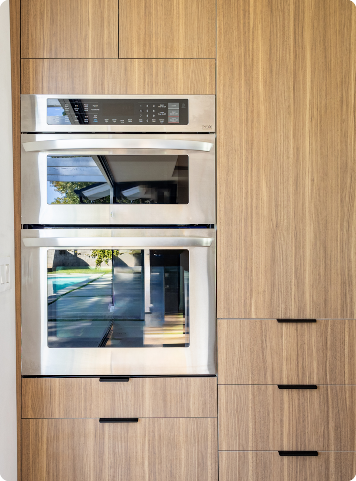 Un four double en acier inoxydable intégré dans des armoires en bois sur mesure, reflétant une vue sur une piscine extérieure moderne et un patio.