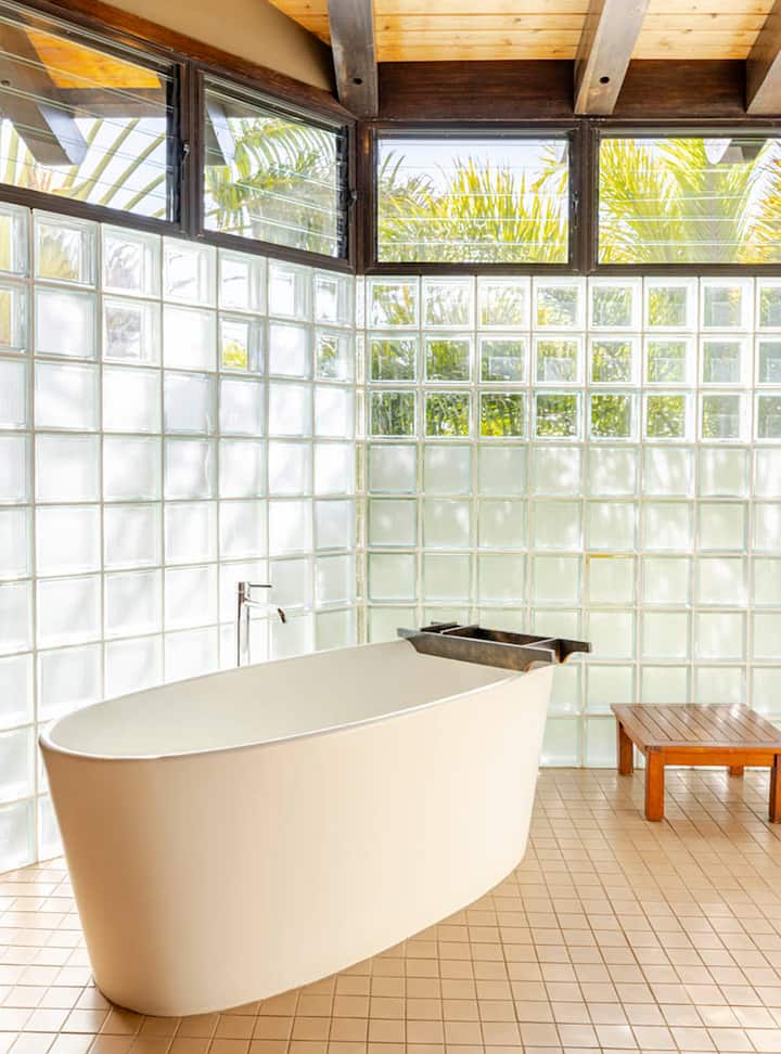 Une salle de bain lumineuse avec une baignoire îlot blanche, des murs en briques de verre, un plafond en bois et de la végétation tropicale visible à travers les fenêtres.