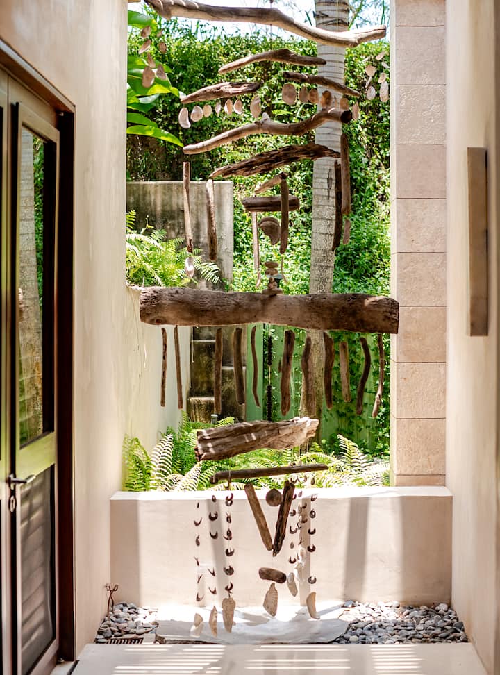 Une installation artistique suspendue composée de bois flotté, de coquillages et d'autres éléments naturels, exposée dans une cour en plein air où la végétation luxuriante laisse passer la lumière du soleil.