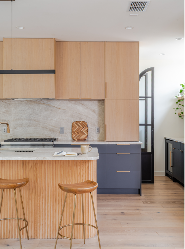 Cuisine moderne avec des placards en bois clair, des placards bas bleu marine, une crédence en marbre et un îlot central avec deux tabourets en bois et un mug sur le comptoir.