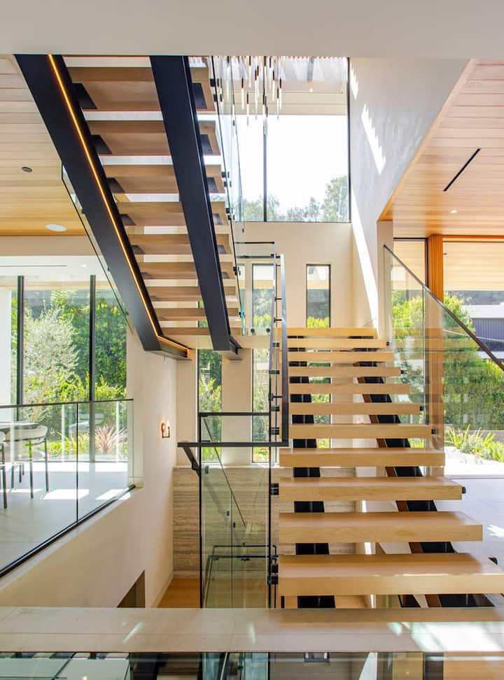 Un escalier ouvert moderne avec des marches en bois et une balustrade en verre, éclairé par la lumière naturelle d'une grande fenêtre donnant sur la verdure.