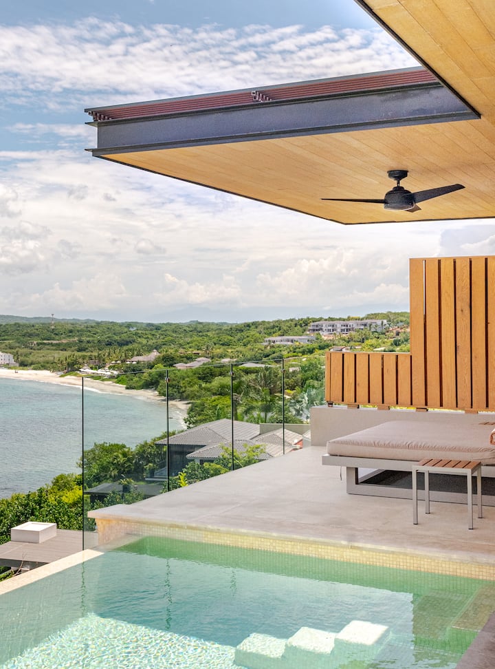 Une piscine à débordement surplombant un paysage verdoyant, une plage de sable et l'océan, avec une architecture moderne en bois et en verre.