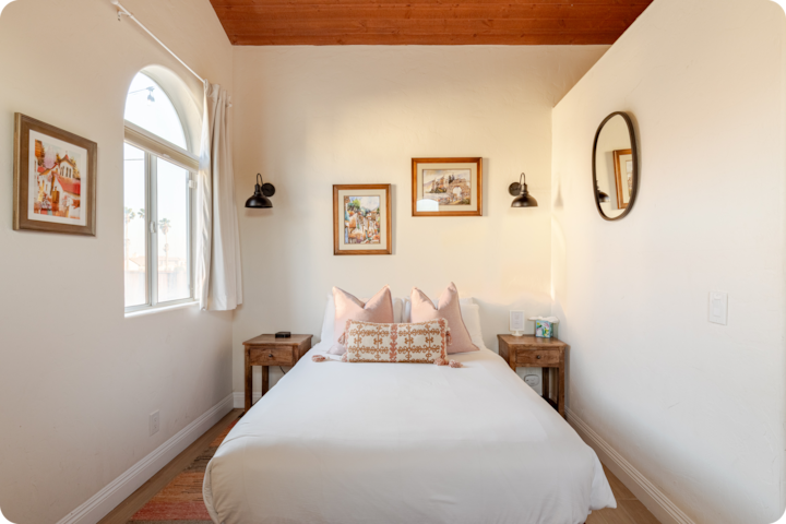 Une chambre confortable avec un lit blanc, des oreillers rose pâle et des coussins décoratifs, encadrée de deux tables de chevet en bois et dotée de deux appliques murales noires assorties. La pièce dispose en outre d'un plafond en bois, d'une fenêtre laissant entrer la lumière naturelle et de tableaux accrochés sur les murs blancs.
