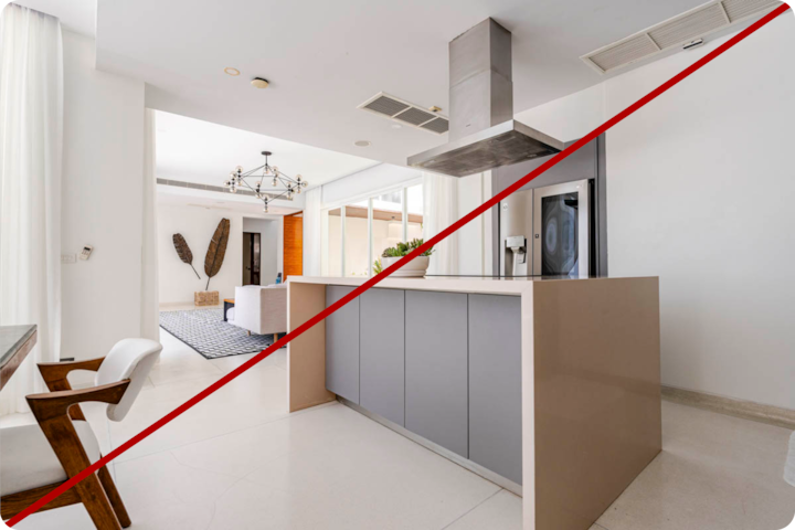 Une cuisine moderne et spacieuse avec un îlot central et une élégante hotte en acier inoxydable, reliée à un salon lumineux doté de grandes fenêtres, d'un lustre et d'un mobilier aux tons neutres. Une ligne diagonale rouge traverse l'image, indiquant une hauteur ou un angle d'appareil photo incorrect.