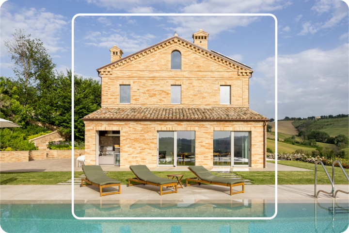 Une vue en plan large d'une villa rustique en briques entourée de collines verdoyantes avec de grandes fenêtres, une terrasse comprenant quatre chaises longues et une piscine au premier plan, sous un ciel bleu vif.