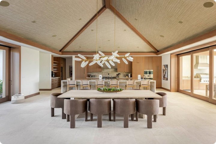 Un coin repas spacieux avec une longue table en bois, dix chaises rembourrées, un lustre moderne et une cuisine ouverte en arrière-plan avec des armoires en bois et des appareils électroménagers en inox.