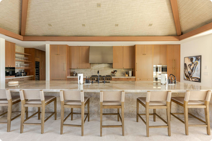 Une cuisine moderne spacieuse avec un grand îlot en marbre, six tabourets de bar en bois, des placards en bois, des appareils électroménagers intégrés et une peinture décorative sur le mur.