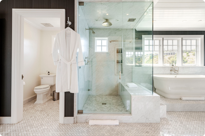 Salle de bain luxueuse avec une douche aux parois vitrées, une baignoire îlot sous de grandes fenêtres avec une vue panoramique et un peignoir blanc suspendu près de l'entrée.