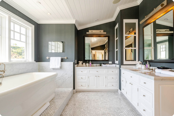 Salle de bain luxueuse avec une baignoire îlot blanche, deux lavabos, des plans vasque en marbre et de grands miroirs reflétant la lumière naturelle des fenêtres donnant sur la verdure.