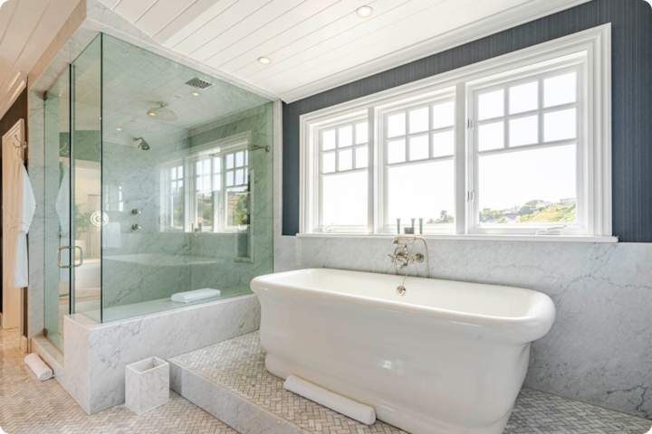 Salle de bain luxueuse avec une baignoire îlot blanche, une douche à l'italienne vitrée et de grandes fenêtres laissant entrer la lumière naturelle et offrant une vue sur la végétation extérieure.