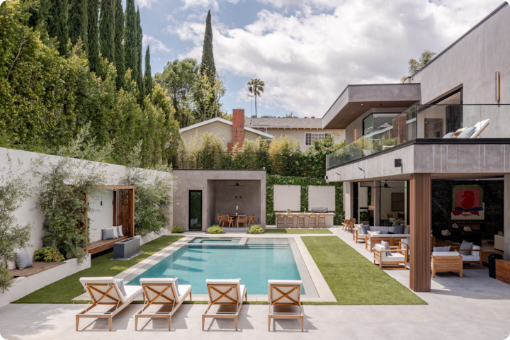 Une cour moderne avec une piscine rectangulaire entourée de chaises longues, un coin salon ombragé et une végétation luxuriante, où se trouve aussi en arrière-plan une maison sur deux niveaux dotée de grandes baies vitrées.