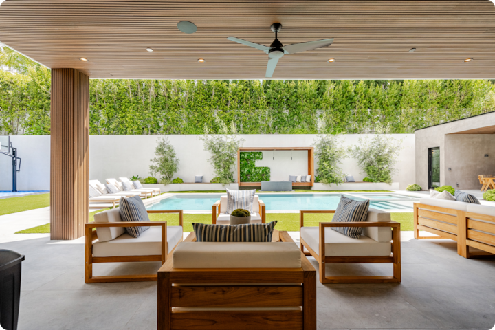 Patio couvert avec salon en bois donnant sur une piscine moderne avec espace détente et entourée de verdure.