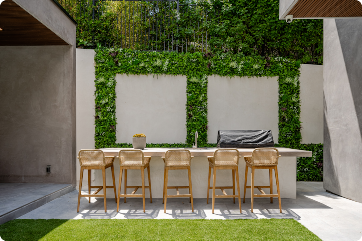 Patio avec un long comptoir entouré de six tabourets en osier, un évier et un barbecue, avec des plantes suspendues et des murs blancs en arrière-plan.