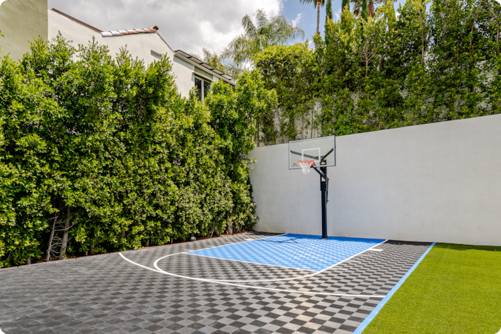 Un terrain de basket-ball extérieur au sol en damiers bleus et noirs, entouré de hautes haies vertes et d'un mur blanc, à côté d'une maison sous un ciel partiellement nuageux.