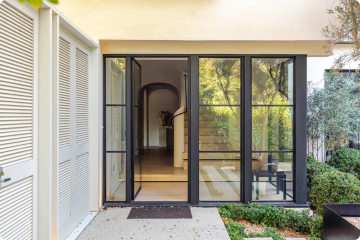 Une entrée moderne en verre avec des portes et des fenêtres au cadre noir, menant à un intérieur lumineux avec un escalier en colimaçon, entourée d'une végétation luxuriante.