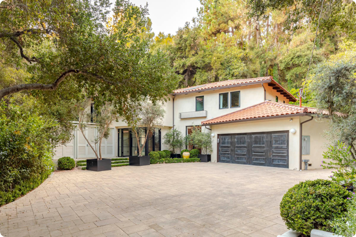 Une maison de style méditerranéen avec un toit de tuiles rouges et une grande allée, entourée d'une végétation et d'arbres luxuriants.