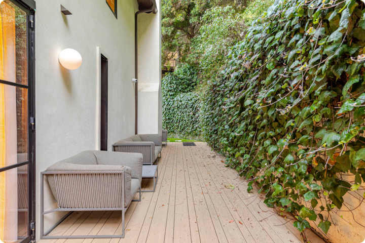 Une terrasse extérieure en bois avec deux fauteuils gris et un mur de lierre vert luxuriant à côté d'un bâtiment blanc moderne.