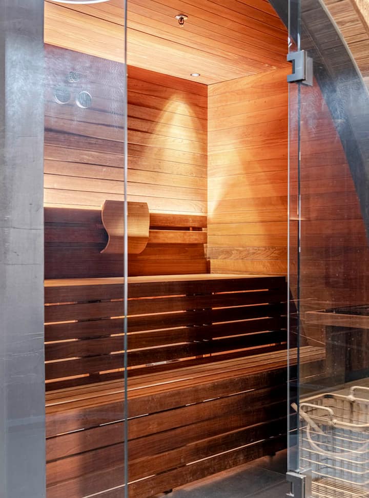 Un sauna moderne en bois avec des portes vitrées, doté d'un éclairage chaleureux, d'un banc en bois et d'un panier dans le coin.