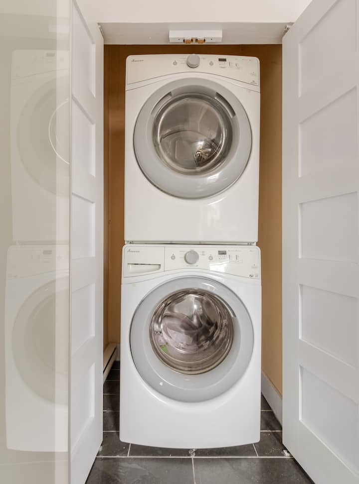 Un lave-linge et un sèche-linge blancs empilés sur le sol carrelé d'un petit placard à linge aux portes pliantes blanches.