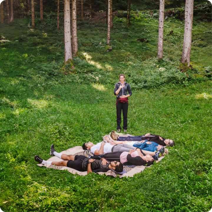 Une guide mène une expérience de méditation dans la nature, dans une clairière au milieu d'une forêt luxuriante. Des personnes sont allongées sur une couverture posée sur l'herbe, détendues et les yeux fermés.