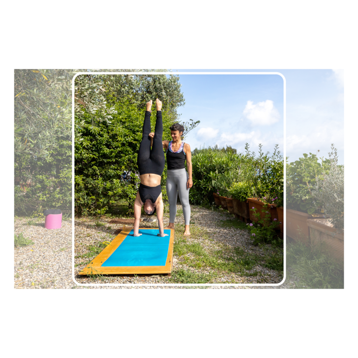 Une personne s'entraîne à faire le poirier sur un tapis de yoga bleu en extérieur, aidée par une instructrice dans un jardin luxuriant sous un ciel bleu.