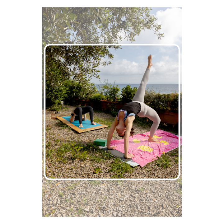 Deux personnes pratiquent le yoga en plein air sur des tapis. L'une d'elles est dans une posture de flexion arrière et l'autre dans la posture de l'enfant. Elles sont entourées de verdure et ont une vue panoramique sur l'océan.