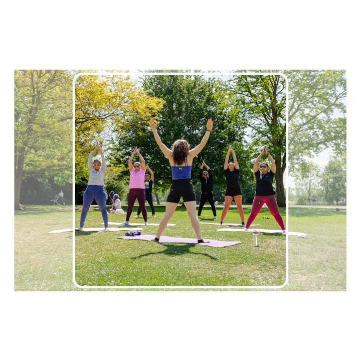 Une professeure de yoga dirige un groupe de cinq personnes en posture de yoga debout sur des tapis dans un parc ensoleillé. Ils sont entourés d'arbres verts.