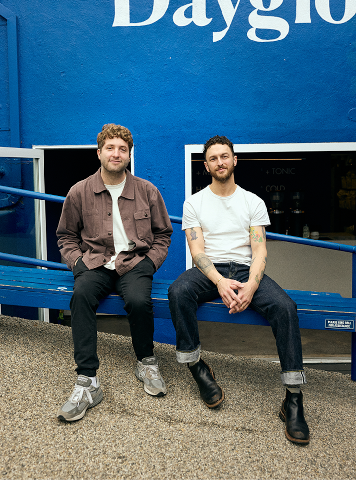 Deux personnes sont assises sur un banc bleu. L'une d'elles porte une veste marron et des baskets, l'autre une chemise blanche, des tatouages et des bottes noires. En arrière-plan, un mur bleu vif présente l'inscription « Dayglo ».