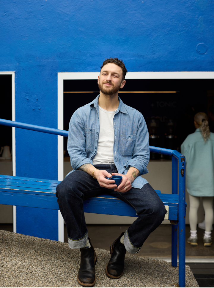 Une personne est assise sur un banc bleu vif et tient une petite tasse bleue. Un mur bleu vif et l'entrée d'un café sont visibles en arrière-plan.