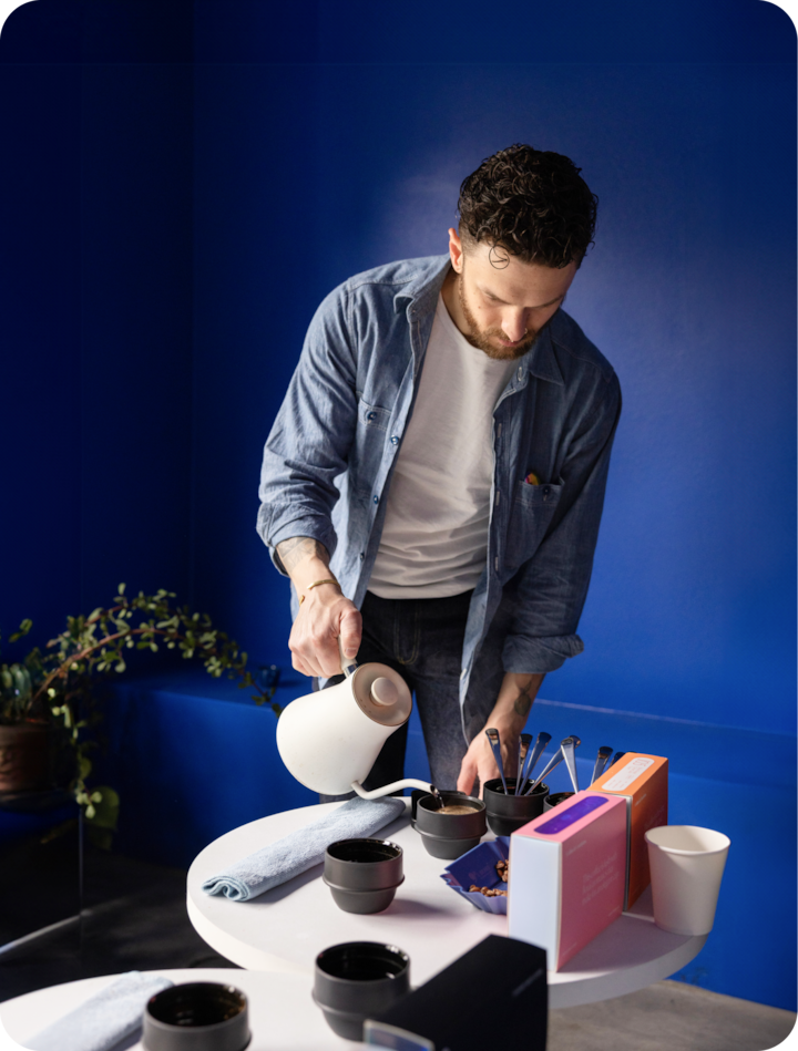 Une personne verse de l'eau chaude à l'aide d'une bouilloire blanche dans une tasse noire posée sur une petite table avec des ustensiles permettant de préparer le café, un torchon et des emballages colorés, devant un mur bleu vif.