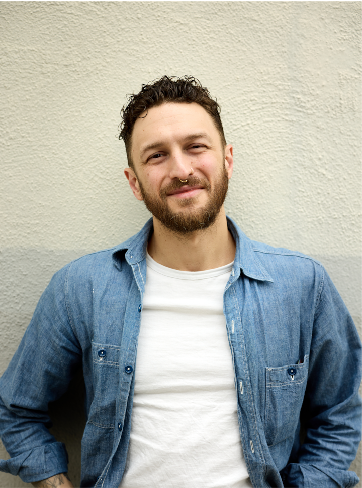 Gros plan d'une personne portant un t-shirt blanc décontracté et une chemise en jean ouverte, debout contre un mur de couleur neutre.