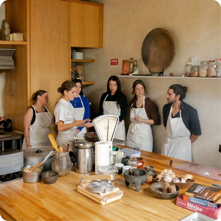 Un groupe de personnes portant des tabliers se tient autour d'une table en bois dans une cuisine rustique, écoutant un enseignant pendant un cours de cuisine. La table est couverte d'ustensiles de cuisine, d'ingrédients et de livres de cuisine, dont un intitulé « Mexico ».