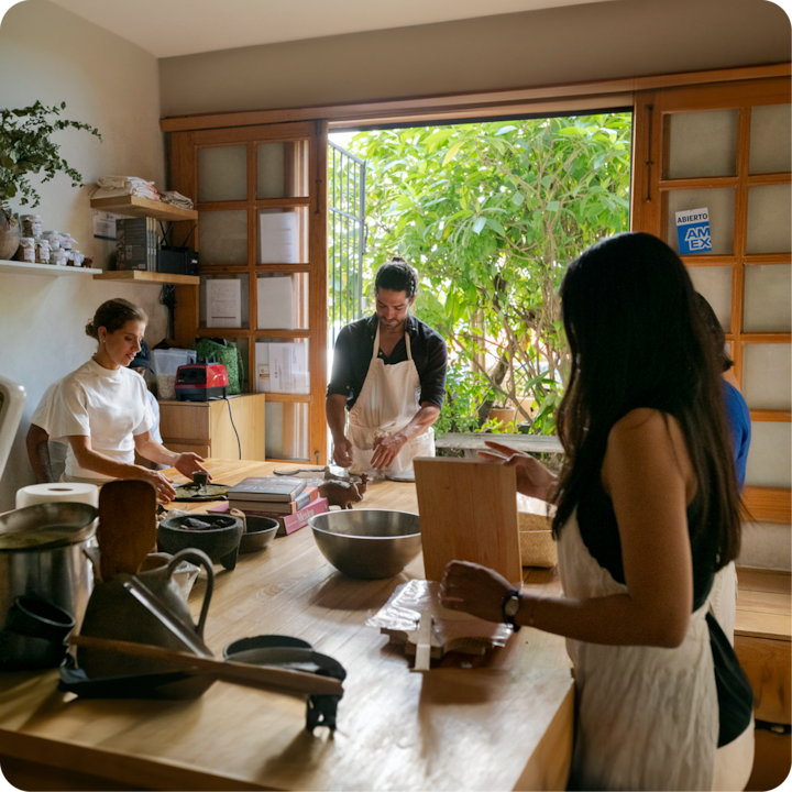 Un cours de cuisine avec des participants portant des tabliers, rassemblés autour d'une table avec des saladiers, des ustensiles et des ingrédients, dans une cuisine ensoleillée. On peut apercevoir de la verdure à l'extérieur.
